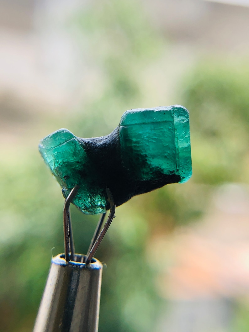 May include: A dark green emerald gemstone with a rough, faceted surface. The emerald is held in place by a pair of metal tweezers.