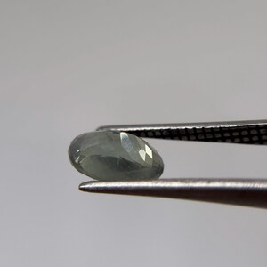 May include: A faceted, pear-shaped gemstone in shades of pale green and gray is held by a pair of tweezers. The gem has multiple facets that reflect light, creating a sparkling effect. The tweezers are silver-colored.