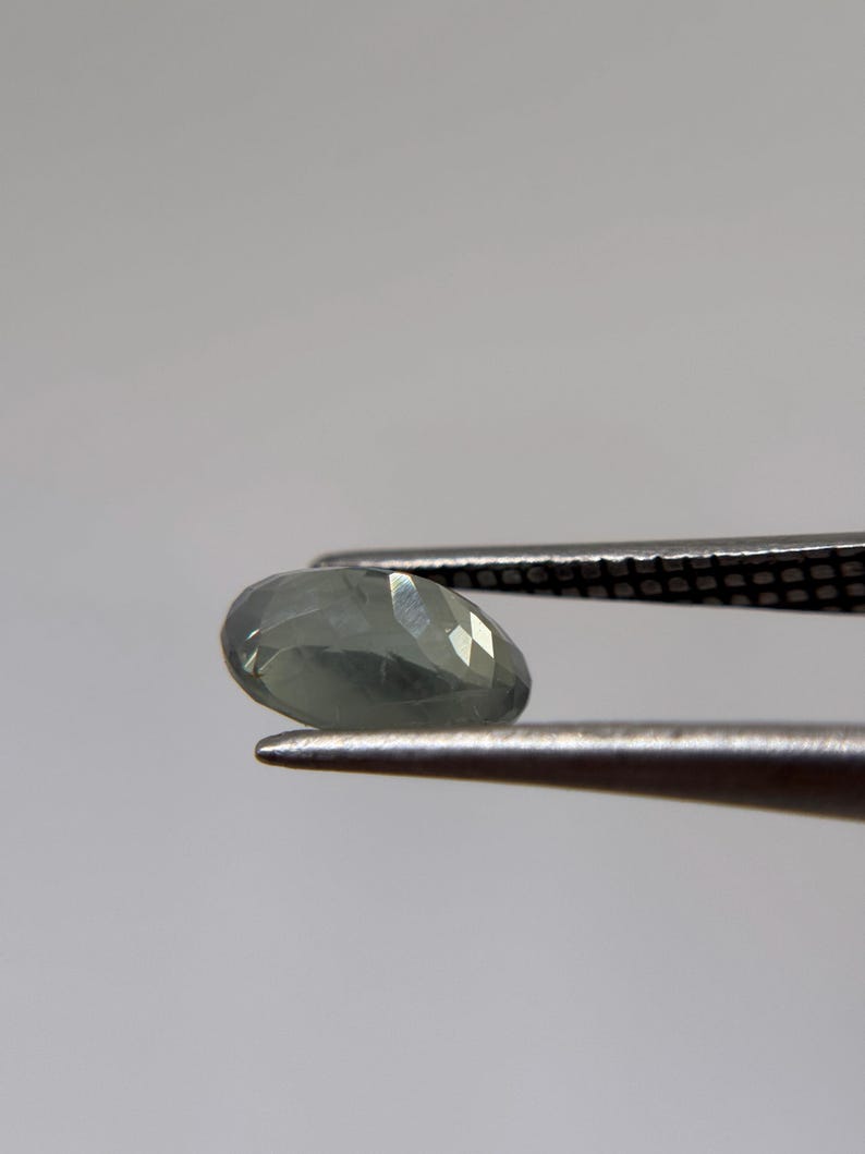 May include: A faceted, pear-shaped gemstone in shades of pale green and gray is held by a pair of tweezers. The gem has multiple facets that reflect light, creating a sparkling effect. The tweezers are silver-colored.