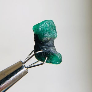 May include: A rough, green emerald gemstone with black inclusions, held by a pair of tweezers.