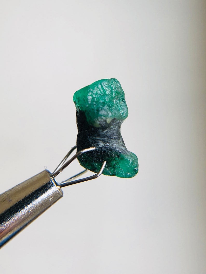 May include: A rough, green emerald gemstone with black inclusions, held by a pair of tweezers.