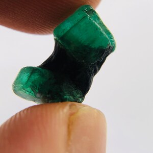 May include: A dark green emerald crystal with a rough, natural texture. The crystal is shaped like a small, irregular block.