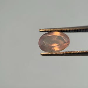 May include: A faceted, oval-shaped gemstone held by tweezers. The gem has a peach and light orange color with a translucent appearance. The tweezers are a dark metal, and the background is a light gray.