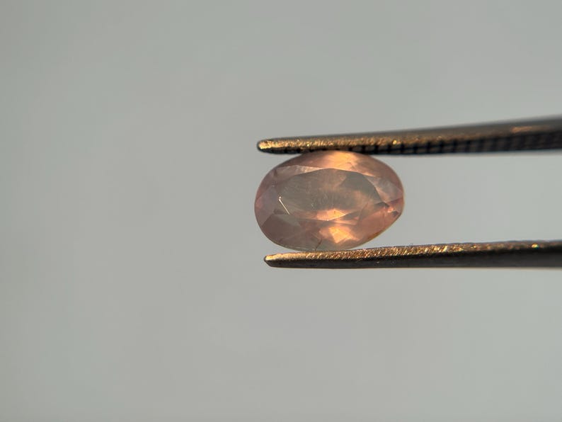 May include: A faceted, oval-shaped gemstone held by tweezers. The gem has a peach and light orange color with a translucent appearance. The tweezers are a dark metal, and the background is a light gray.