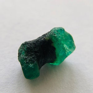 May include: A rough, green emerald gemstone with a dark green inclusion. The emerald has a natural, unpolished look.