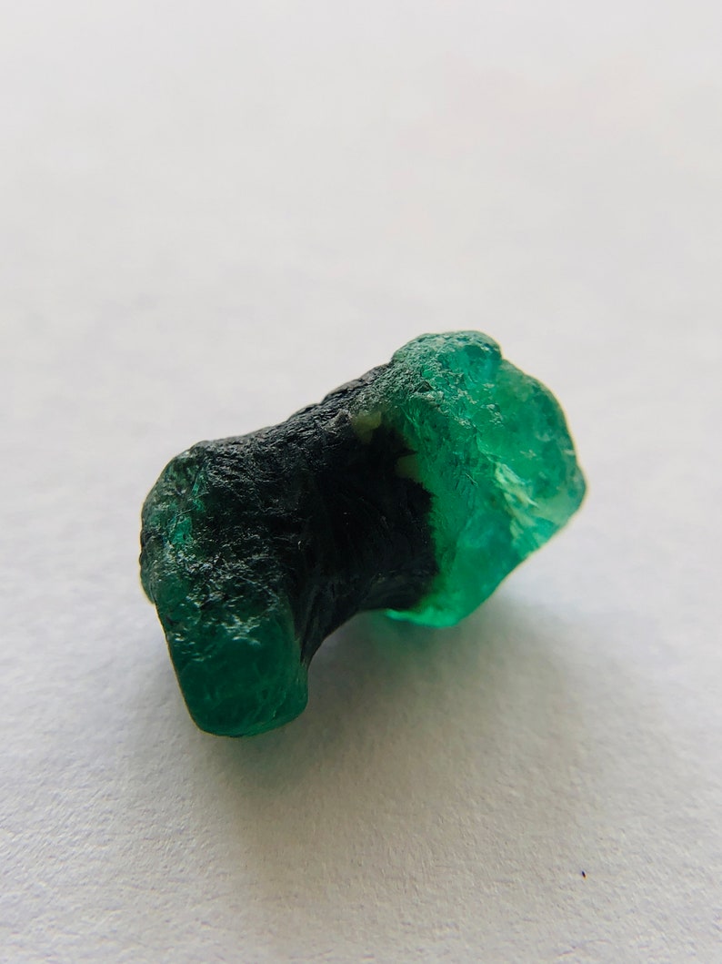 May include: A rough, green emerald gemstone with a dark green inclusion. The emerald has a natural, unpolished look.