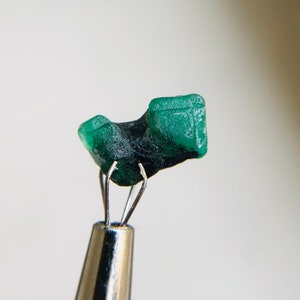 May include: A vibrant green emerald crystal, with a unique shape, is held by a pair of tweezers. The crystal has a smooth, polished surface and is a beautiful example of this gemstone.