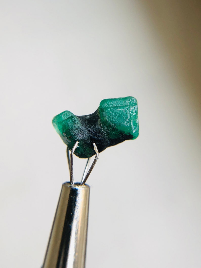 May include: A vibrant green emerald crystal, with a unique shape, is held by a pair of tweezers. The crystal has a smooth, polished surface and is a beautiful example of this gemstone.
