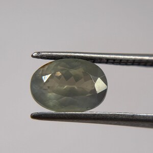 May include: A faceted, oval-shaped gemstone in shades of green and gray is held by a pair of tweezers. The gem has multiple facets that reflect light, creating a sparkling effect. The background is a plain, light gray.