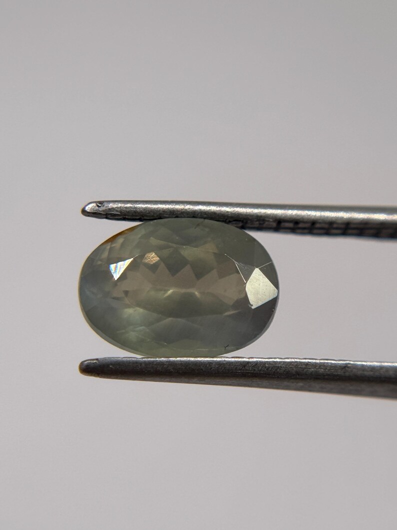 May include: A faceted, oval-shaped gemstone in shades of green and gray is held by a pair of tweezers. The gem has multiple facets that reflect light, creating a sparkling effect. The background is a plain, light gray.