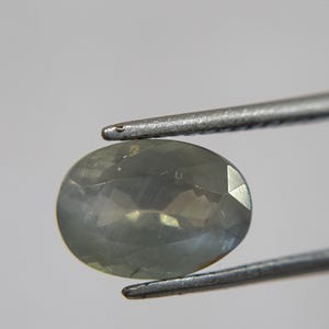May include: An oval-shaped, pale green gemstone is held by a pair of silver tweezers. The gemstone has a faceted surface that reflects light, creating a subtle sparkle. The background is a soft, neutral gray.