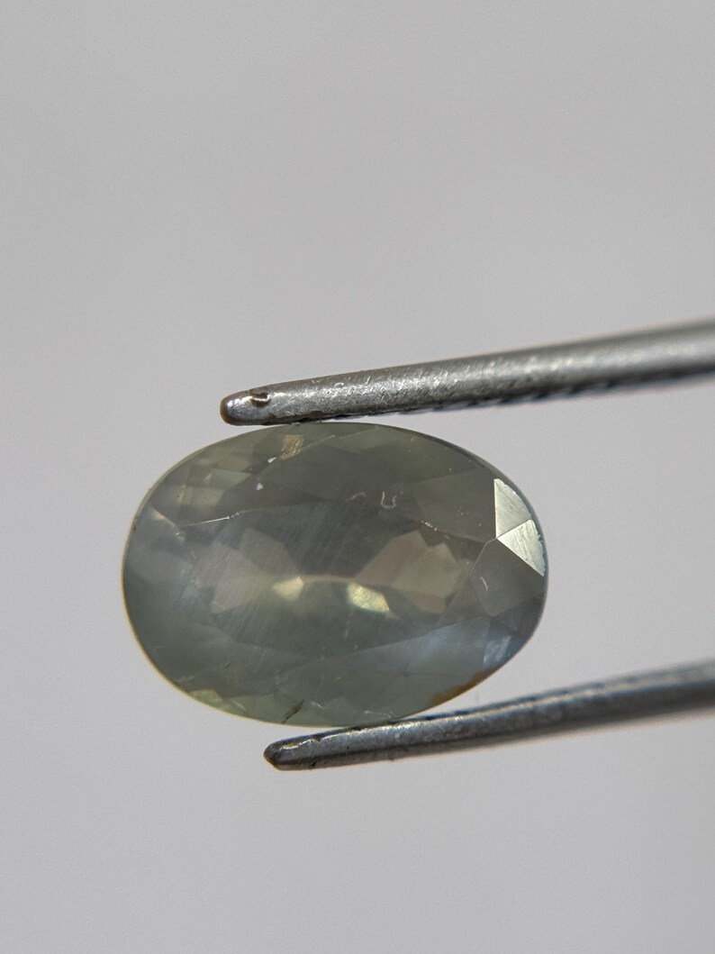 May include: An oval-shaped, pale green gemstone is held by a pair of silver tweezers. The gemstone has a faceted surface that reflects light, creating a subtle sparkle. The background is a soft, neutral gray.