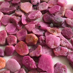 May include: A collection of pink ruby gemstone chips. The chips are various sizes and shapes, with some having a more reddish hue. The chips are scattered on a white surface.