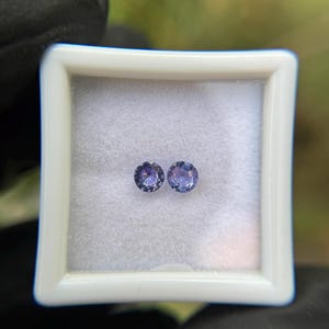 May include: Two round, purple gemstones in a white box. The gemstones are approximately 5mm in diameter.