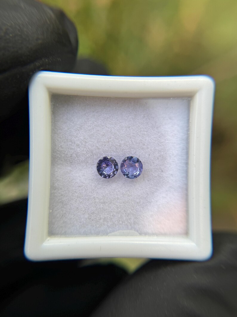 May include: Two round, purple gemstones in a white box. The gemstones are approximately 5mm in diameter.