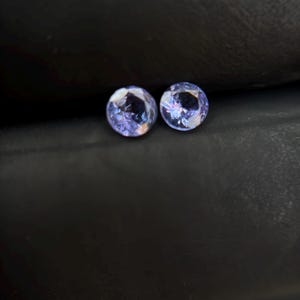 May include: Two round, purple gemstones with a light blue hue, set against a dark gray background.