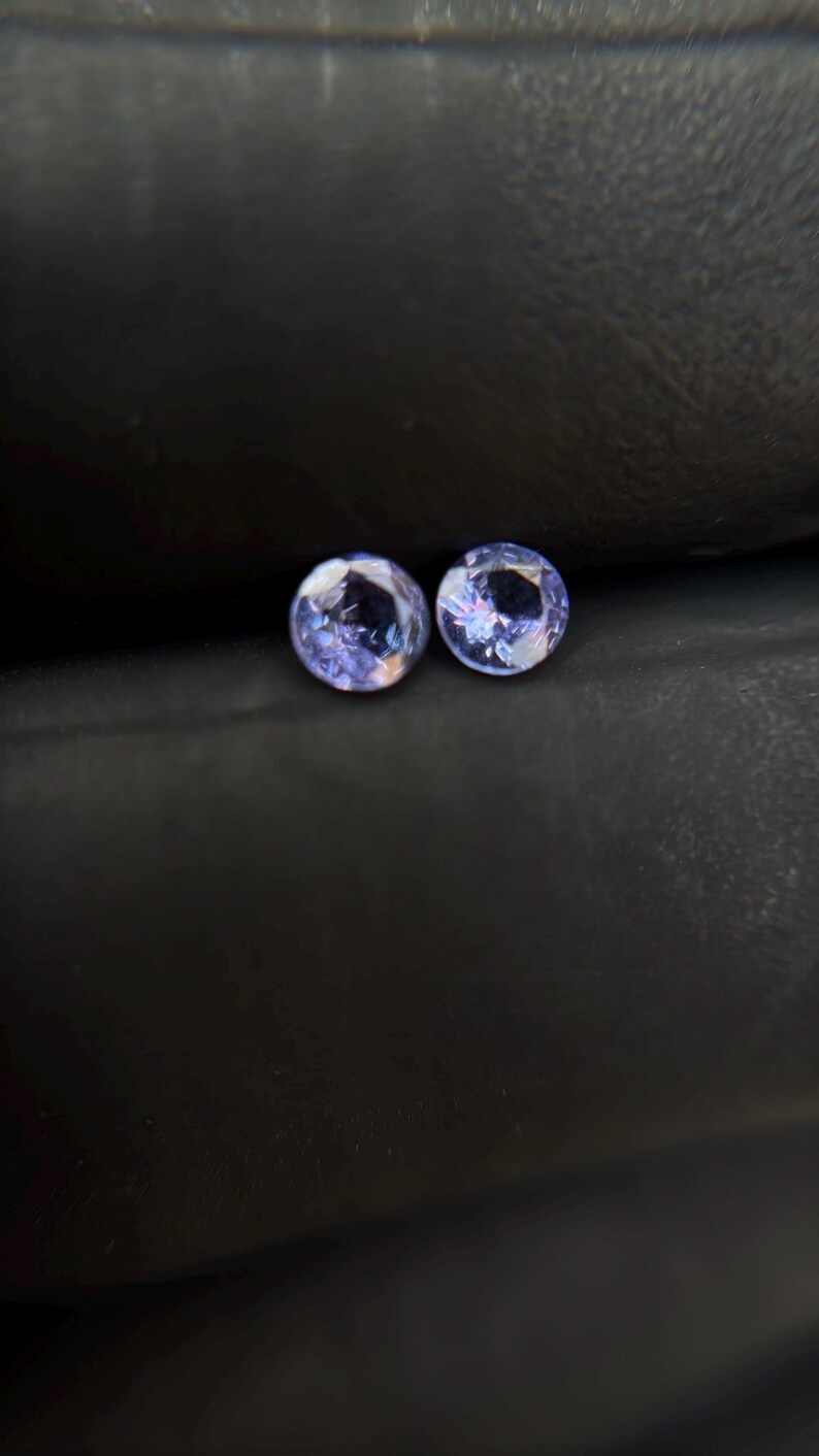 May include: Two round, purple gemstones with a light blue hue, set against a dark gray background.