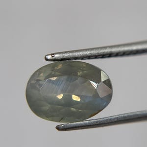 May include: An oval-shaped, pale green gemstone is held by a pair of silver tweezers. The gem has multiple facets that reflect light, creating a shimmering effect. The background is a plain, light gray.