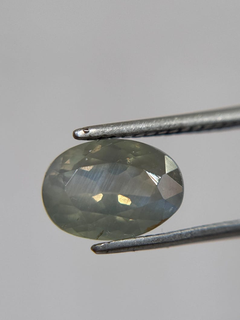 May include: An oval-shaped, pale green gemstone is held by a pair of silver tweezers. The gem has multiple facets that reflect light, creating a shimmering effect. The background is a plain, light gray.