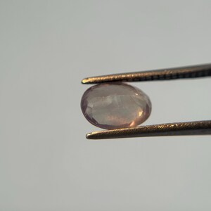 May include: A light pink, oval-shaped gemstone is held by a pair of tweezers. The gem has a translucent quality, with subtle internal patterns. The tweezers are dark metal, contrasting with the gem's soft color. The background is a plain, light gray.