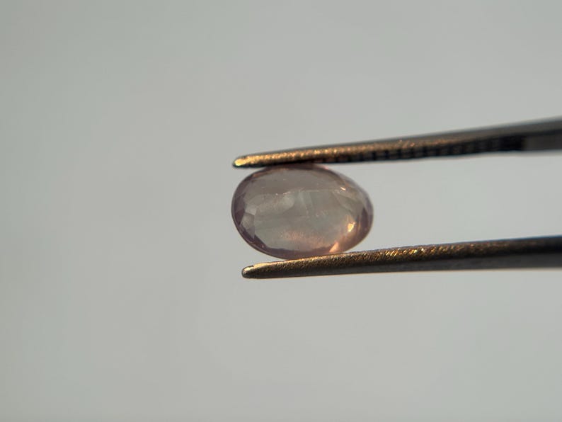 May include: A light pink, oval-shaped gemstone is held by a pair of tweezers. The gem has a translucent quality, with subtle internal patterns. The tweezers are dark metal, contrasting with the gem's soft color. The background is a plain, light gray.