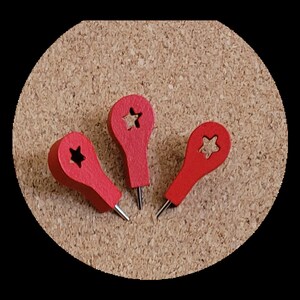 May include: Three red push pins with a star cut out in the centre of each pin head. The push pins are on a cork board.