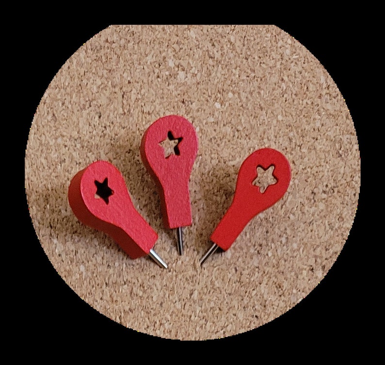 May include: Three red push pins with a star cut out in the centre of each pin head. The push pins are on a cork board.