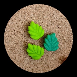 May include: Three decorative leaf-shaped pins, two are green and one is teal, on a corkboard background.