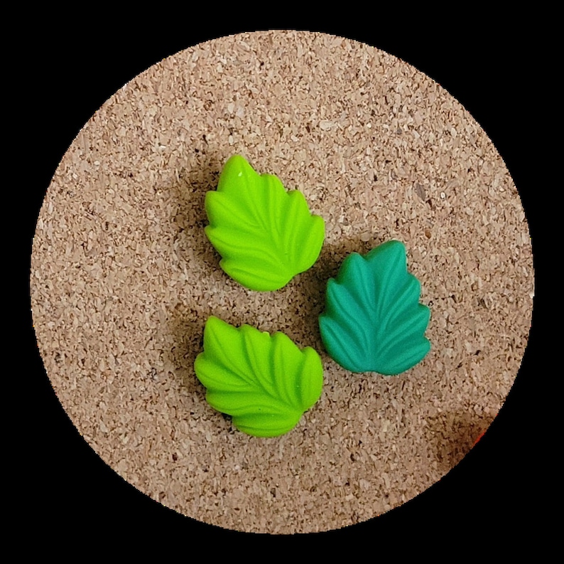 May include: Three decorative leaf-shaped pins, two are green and one is teal, on a corkboard background.