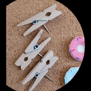 May include: Three wooden clothes pegs with metal springs are pinned to a corkboard. Two pink and blue donut-shaped decorations are also pinned to the board.