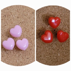 May include: Three pink heart-shaped push pins and three red heart-shaped push pins on a corkboard background.