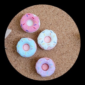 May include: Four colourful donut-shaped push pins with sprinkles. The donuts are pink, blue, and purple with white frosting.