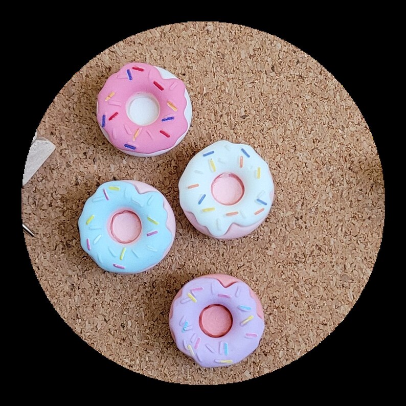 May include: Four colourful donut-shaped push pins with sprinkles. The donuts are pink, blue, and purple with white frosting.