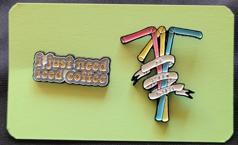 May include: Two enamel pins. One pin says "I just need iced coffee" in a retro font. The other pin features three colorful straws with a white banner that says "No Straw Please".