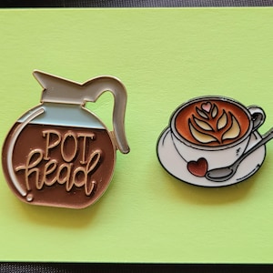 May include: Two enamel pins, one featuring a coffee pot with the text "POT head" and the other a cup of coffee with latte art.
