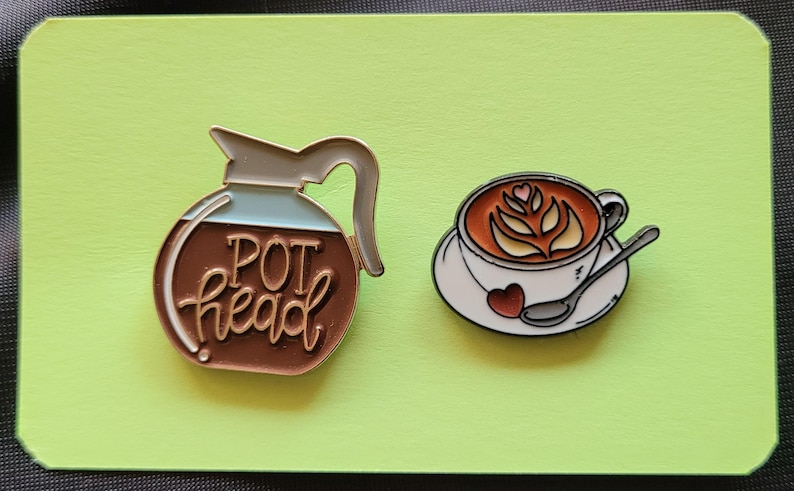 May include: Two enamel pins, one featuring a coffee pot with the text "POT head" and the other a cup of coffee with latte art.