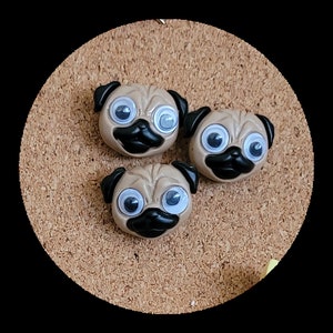 May include: Three brown pug-shaped push pins with googly eyes and black noses. The push pins are on a corkboard.