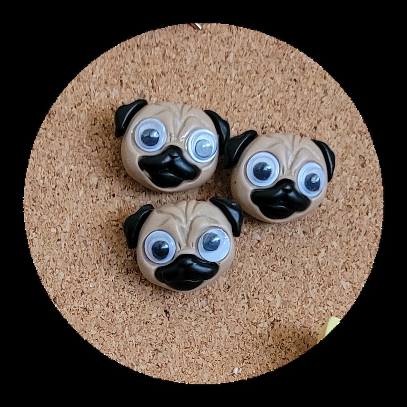 May include: Three brown pug-shaped push pins with googly eyes and black noses. The push pins are on a corkboard.