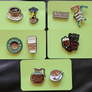 May include: Set of ten enamel pins with coffee themes. The pins feature images of coffee cups, coffee pots, and coffee-related sayings. The pins are on a green background.