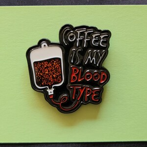 May include: Black and red enamel pin with a coffee bag and the text "Coffee is my blood type".