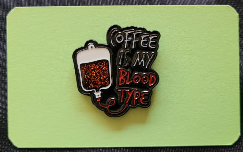 May include: Black and red enamel pin with a coffee bag and the text "Coffee is my blood type".