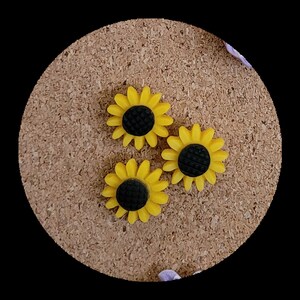 May include: Three yellow sunflower-shaped push pins with black centres. The push pins are on a corkboard.