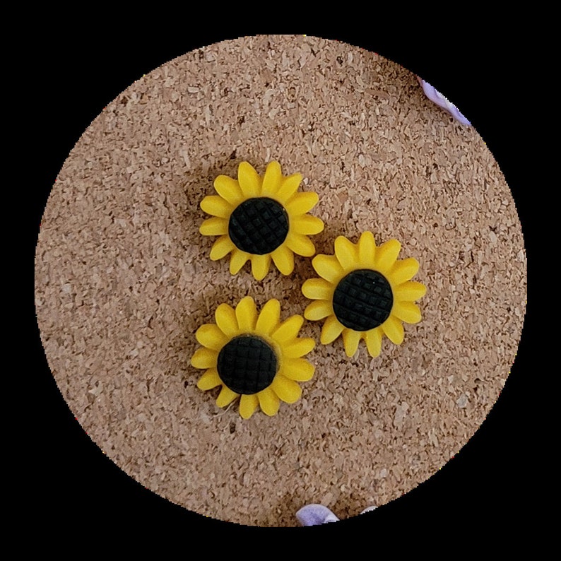 May include: Three yellow sunflower-shaped push pins with black centres. The push pins are on a corkboard.
