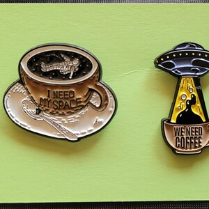May include: Two enamel pins featuring coffee mugs with space-themed designs. The first pin shows an astronaut floating in a mug of coffee with the text "I need my space". The second pin shows a UFO pouring coffee into a mug with the text "We need coffee".