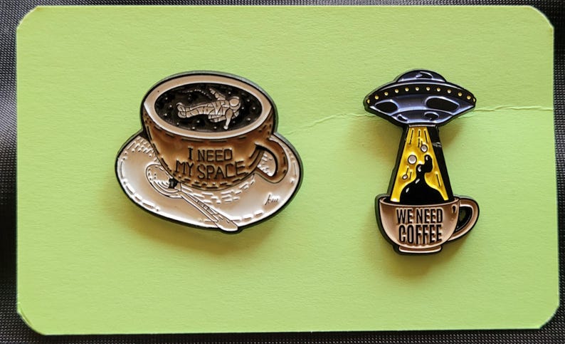 May include: Two enamel pins featuring coffee mugs with space-themed designs. The first pin shows an astronaut floating in a mug of coffee with the text "I need my space". The second pin shows a UFO pouring coffee into a mug with the text "We need coffee".