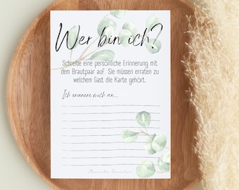 May include: A white card with green watercolor leaves and the text "Wer bin ich?"  The card has lines for writing and the text "Schreibe eine persönliche Erinnerung mit dem Brautpaar auf. Sie müssen erraten zu welchem Gast die Karte gehört. Ich erinnere mich an..."  The card is on a wooden surface.