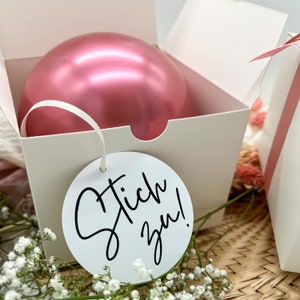 May include: A white gift box with a pink balloon inside. The box has a white tag attached to it that says "Tick you!" in black cursive writing.