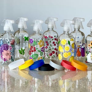 May include: A collection of clear glass spray bottles with various decorative designs, including paw prints, flowers, cherries, lemons, and sunflowers. Each bottle has a white spray nozzle. Colorful silicone bands are in the foreground.