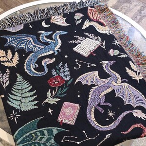 May include: A black woven throw blanket featuring a multi-colored pattern of dragons, open books, ferns, flowers, and constellations. The blanket has a fringed edge with multi-colored threads along the top and side borders of the fabric.
