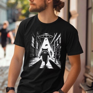 May include: Black t-shirt featuring a white graphic of a raccoon running down a city street with a UFO above. The raccoon is being chased by two aliens.